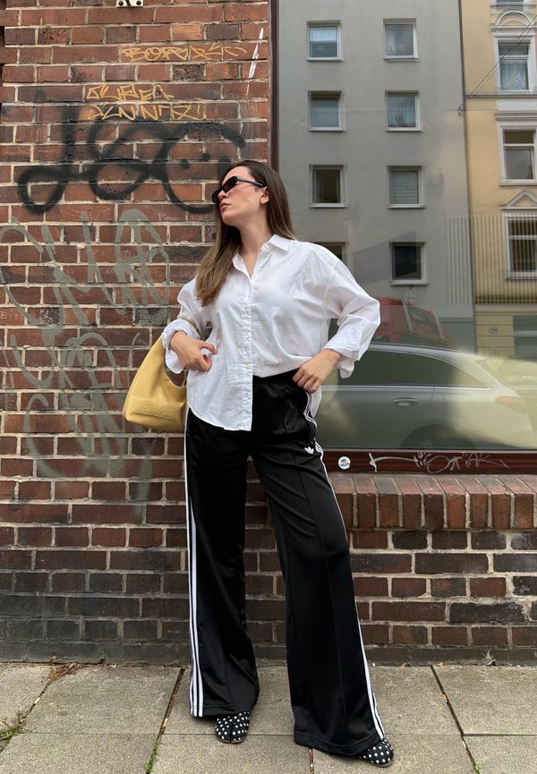 Nainen valkoisessa paidassa, mustissa valkoisilla raidoilla varustetuissa Adidas-housuissa ja aurinkolaseissa poseeraa tiiliseinää vasten, jossa on graffitia ja ikkuna heijastaa rakennuksia.