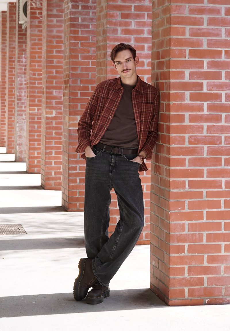 Joven con camisa de cuadros marrones, jeans oscuros y botas apoyado contra una columna de ladrillo rojo en un pasillo iluminado por el sol con arcos.