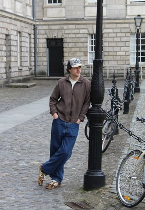Chaqueta de punto marrón con cremallera, camisa crema, vaqueros azul desgastado holgados y zapatillas marrones con patrones. Un hombre se apoya en un poste de luz en una zona de adoquines.