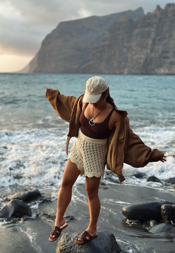 Kvinde i beige kasket, brun top og strikkede shorts, der balancerer på sten ved havet med store klipper i baggrunden under solnedgang.