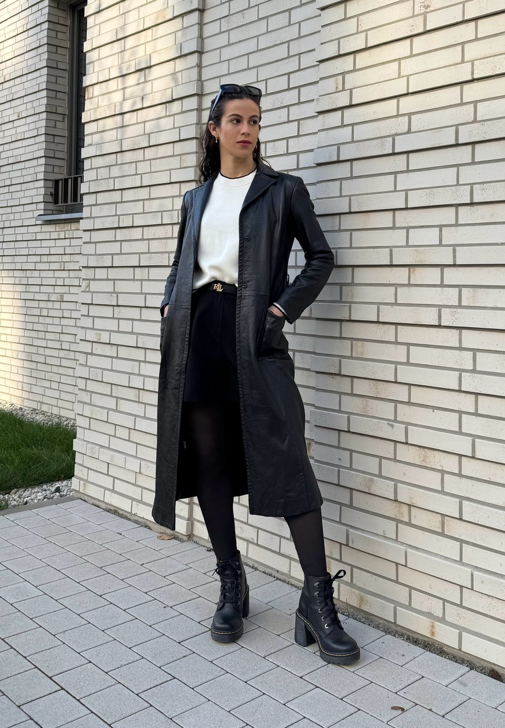 Čierny kožený dlhý kabát so strihom na mieru, nosený cez biely sveter a čiernu sukňu. Čierne šnurovacie podpätkové čižmy celý outfit dopĺňajú.