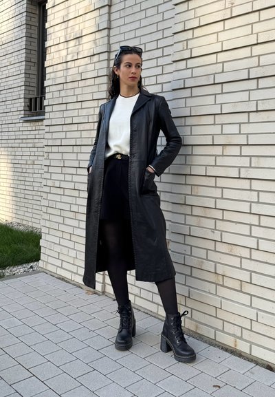 Abrigo largo de cuero negro con un diseño a medida, llevado sobre un suéter blanco y una falda negra. Botines de tacón negro con cordones completan el conjunto.