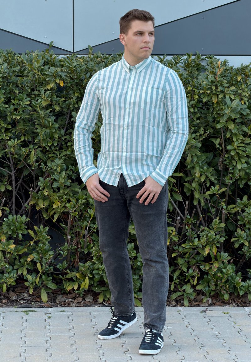 Jauns vīrietis tumši zaļā un baltā svītraina ar pogām jaka, melnās džinsās un melnās sporta kurpēs stāv uz bruģēta celiņa priekšā zaļam dzīvžogam.