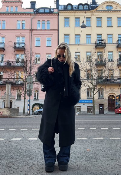 Abrigo largo negro con puños de felpa, superpuesto a una bufanda negra. Combinado con pantalones de pierna ancha azul oscuro y zapatos de plataforma en un entorno urbano.