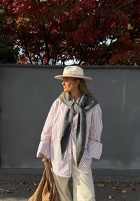 Lys rosa stripete skjorte med vide ermer, grått skjerf knyttet rundt halsen, kremfargede vide bukser og en beige hatt med bred brem. Brun vevd veske.