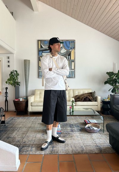 Camisa blanca de manga larga, pantalones cortos negros hasta la rodilla, calcetines blancos, zapatos negros. Entorno interior con muebles modernos y vegetación.