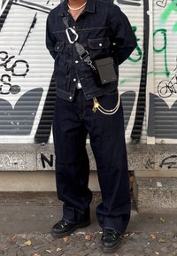 Veste en denim noir avec deux poches poitrine, portée sur un jean noir ample assorti. Sac bandoulière noir, chaînes et baskets texturées.