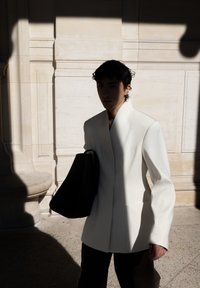 Hvit skreddersydd blazer med høy krage, kombinert med svarte bukser og en stor svart totebag, presentert i skyggefull belysning.