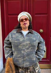 Denim jacket dengan pola kamuflase dan kerah korduroy, dipadukan dengan atasan putih berpola rib dan rok kamuflase; diakses dengan beret krim dan kacamata hitam.