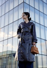 Trench coat in pelle blu scuro con bottoni doppiopetto e cintura. Abbinato a una borsa marrone strutturata. Ambientazione urbana moderna.