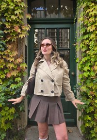 Beige cropped double-breasted jacket, pleated brown skirt, brown handbag, round sunglasses, standing in front of a green door and ivy.