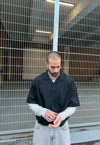 Schwarzes Kurzarmhemd über einem grau-weißen gestreiften Langarmshirt, kombiniert mit hellgrauen Jeans. Die Person trägt eine Kettenkette und Ringe.