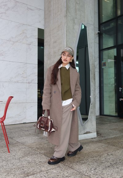 Mujer con abrigo beige, suéter oliva, pantalones beige, zapatos marrones y gorra, de pie junto a un pilar de concreto y una entrada de vidrio, sosteniendo un bolso marrón.