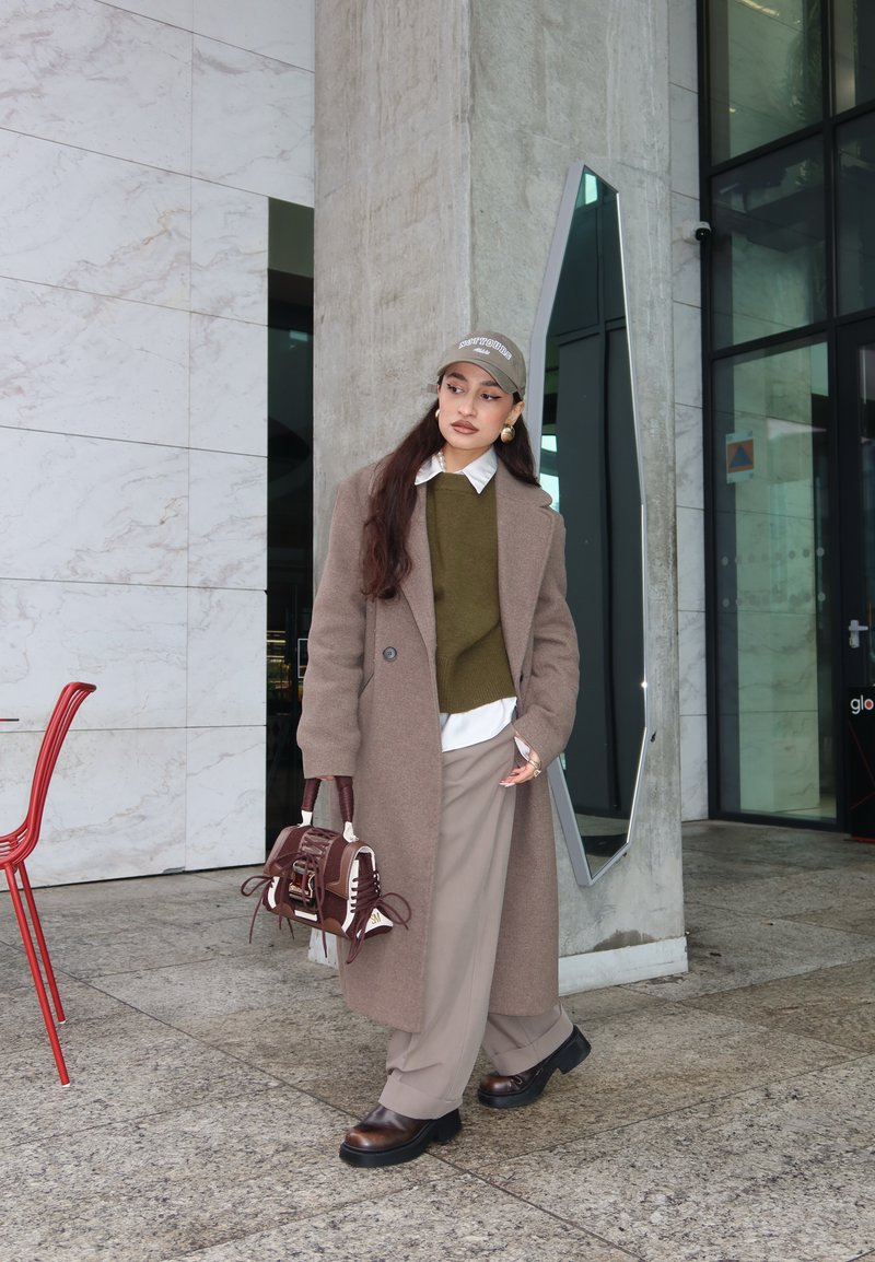 Femme en manteau beige, pull olive, pantalon beige, chaussures marron et casquette se tenant près d'un pilier en béton et d'une entrée en verre, tenant un sac à main marron.