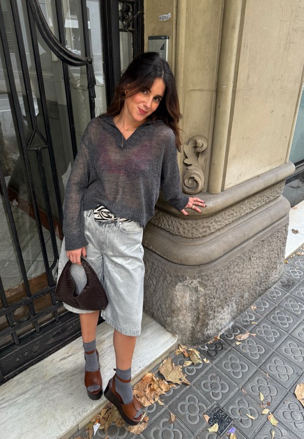 Junge Frau, die sich an der Ecke eines Steinbaus anlehnt. Sie trägt einen grauen Pullover, silberne Shorts, braune Plateausandalen mit grauen Socken und hält eine dunkle Handtasche.