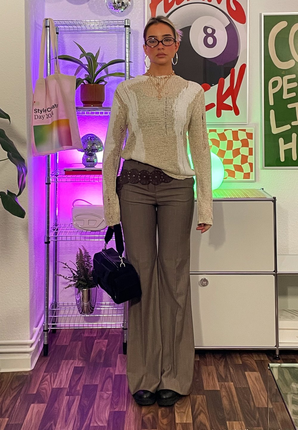 Μπεζ πλεκτό πουλόβερ με υφή, καφέ ψηλόμεσες φαρδιές παντελόνες, μαύρη τσάντα χειρός και στρογγυλά γυαλιά. Εσωτερικός χώρος με φυτά.