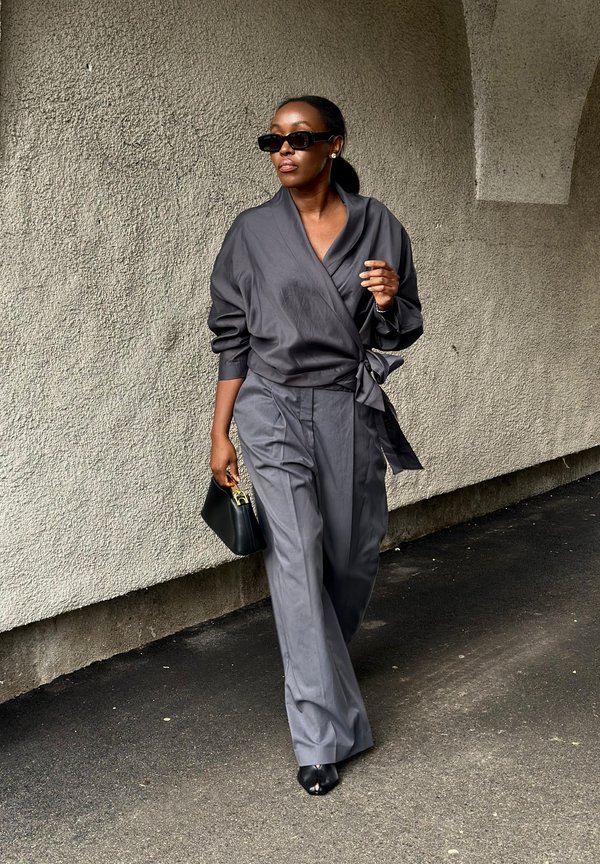 Donna con blusa avvolgente grigia e pantaloni a gamba larga che cammina con una borsa nera e occhiali da sole davanti a una parete di cemento con texture.