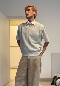 Pulover gri fără mâneci peste o cămașă albă cu guler, asortat cu pantaloni largi maro deschis. Material neted, croială lejeră și design casual.