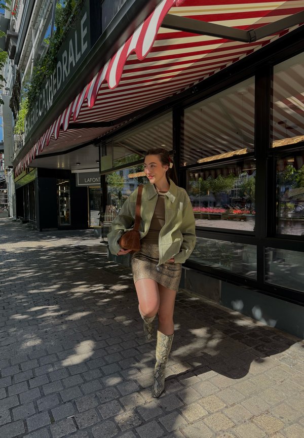 Jonge vrouw met groene jas, rok met slangenprint en laarzen, lopend op een keienstraat naast een café met een rood-wit gestreept zonnescherm.