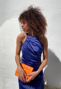 Marineblauwe satijnen jurk met een schouderontwerp en gedrapeerde textuur. Houdt een gequilt oranje clutch vast. De achtergrond heeft een textuur van een witte muur.