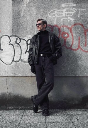 Chaqueta de cuero negra sobre un suéter negro de cuello alto, combinada con unos jeans de pierna ancha oscuros y zapatos negros, frente a una pared cubierta de graffiti.