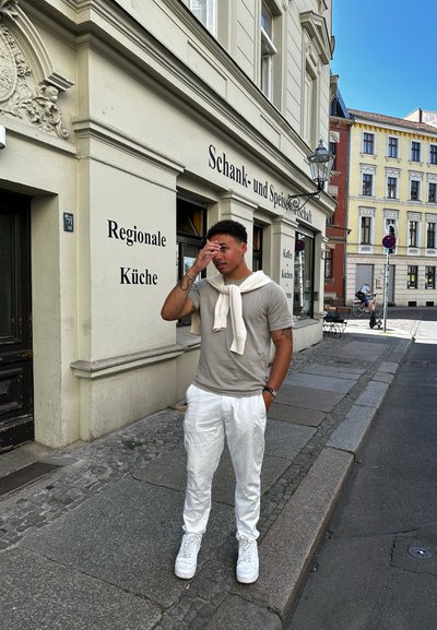 Camiseta gris clara y pantalones blancos con un acabado texturizado. Zapatillas blancas, suéter ligero colgado alrededor del cuello. Entorno urbano.