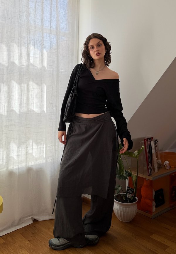 Jeune femme aux cheveux bouclés portant un haut noir épaules dénudées, une jupe portefeuille sombre par-dessus un pantalon large, debout à l'intérieur près de rideaux blancs transparents.