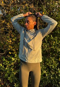 Veste gris clair et légère avec un col montant et une fermeture éclair sur le devant, associée à des leggings vert olive. Cheveux tressés coiffés vers le haut.