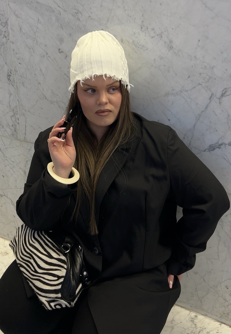 Młoda kobieta w białej dzianinowej czapce i czarnym blezerze, trzymająca okulary przeciwsłoneczne blisko twarzy, z torbą w pasiasty wzór zebry, siedząca przy marmurowej ścianie.