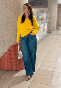 Femme en pull jaune et jeans pattes d'eph appuyée contre une colonne carrelée, tenant un sac à main blanc, debout sur un sol carrelé à l'intérieur.