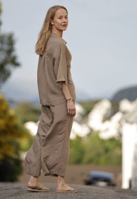 Donna con lunghi capelli biondi che indossa una camicia e pantaloni marroni larghi, con infradito, in piedi all'aperto su una strada con sfondo sfocato.
