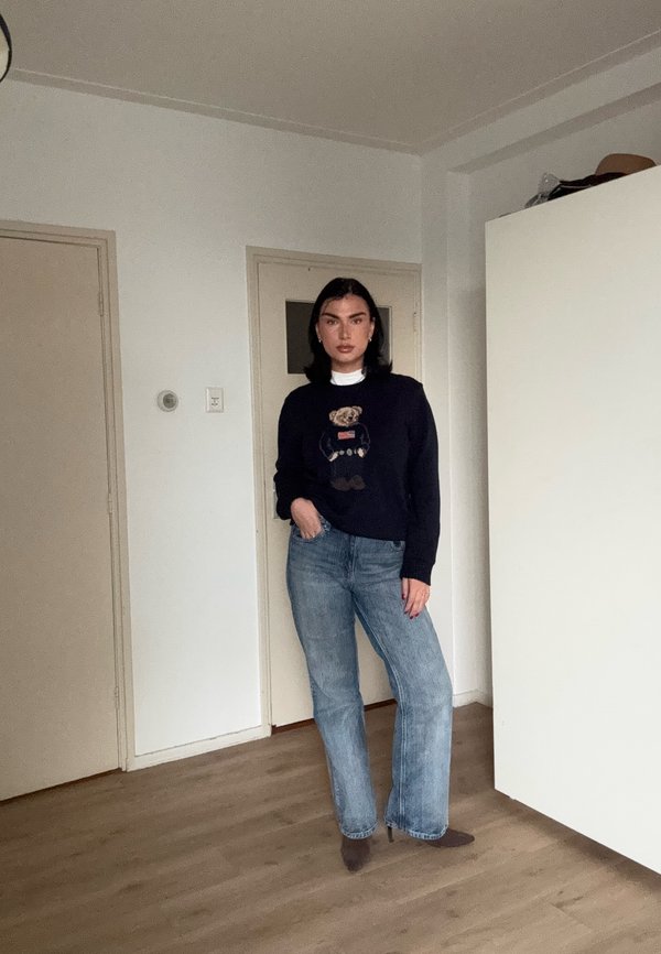 Mujer de pie en el interior con un suéter negro con diseño de oso de peluche, vaqueros azules y zapatos marrones de tacón, posando con una mano en el bolsillo.