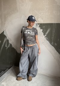 Chemise camouflage grise à manches courtes avec texte blanc, pantalon de jogging gris ample avec rayures, et chaussures marron. Le modèle pose avec un sac gris.