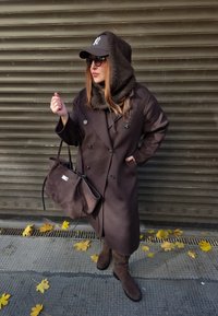 Barna, dupla soros kabát nagy gombokkal, hozzáillő sállal és kézitáskával, fekete sapkával, napszemüveggel és térdig érő barna csizmával, struktúrált háttér előtt.