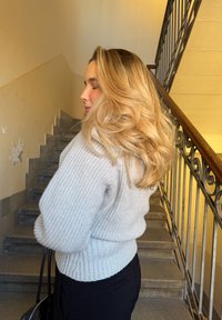 Cardigan en laine gris clair avec un motif tricoté texturé, taille cintrée et poignets côtelés. Le mannequin se tient près d'un escalier, tenant un sac.