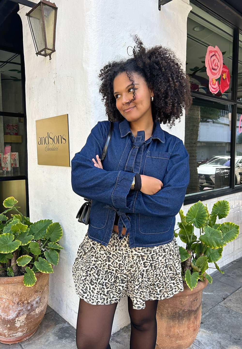 Junge Frau mit lockigem Haar, die eine denimbekleidete Jacke und einen Leopardenmusterrock trägt, steht mit verschränkten Armen vor einem Geschäft mit Topfpflanzen.