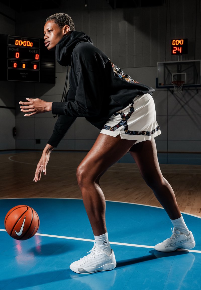 Giocatore di basket con felpa nera e pantaloncini bianchi mentre palleggia un pallone da basket su un campo indoor blu con tabellone segnali e canestro visibili.