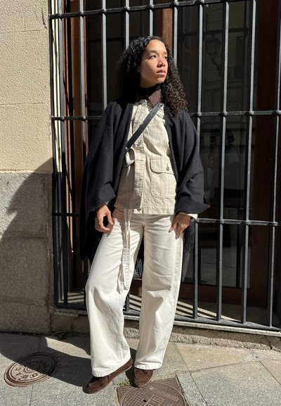 Mujer con cabello rizado vistiendo chaqueta beige, pantalones anchos blancos, chal negro y zapatos marrones, de pie contra barras de hierro en una calle soleada.
