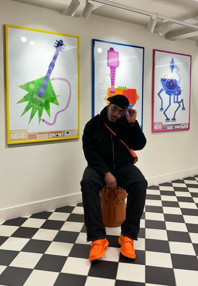 Hombre con atuendo negro y zapatos naranjas sentado en un taburete naranja frente a tres coloridos carteles enmarcados de temática musical abstracta.