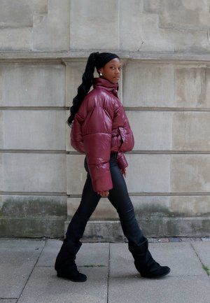 Mujer caminando al aire libre con una brillante chaqueta de plumas burdeos, pantalones negros y botas negras, frente a un fondo de pared de piedra.