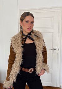 Giovane donna con capelli intrecciati che indossa grandi orecchini a cerchio, un top nero, un giaccone di shearling marrone, una sciarpa nera annodata al collo e una cintura marrone con fibbia decorativa.