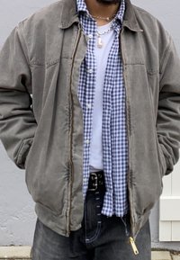 Személy szürke cipzáras dzsekit visel, alatta kék és fehér kockás ing és fehér póló található, fekete farmernadrággal és ezüst nyakláncokkal.