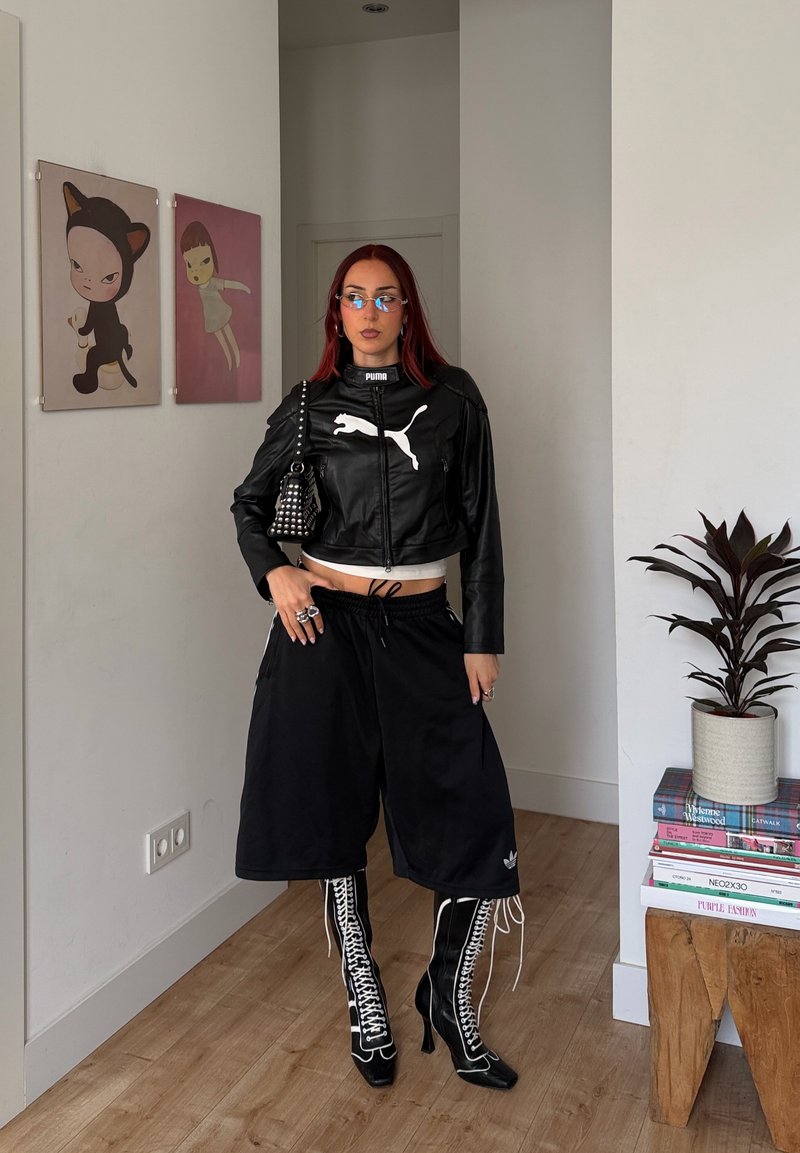 Woman with red hair wearing black Puma jacket, wide black shorts, lace-up high-heeled boots, standing indoors near artwork and a plant on books.