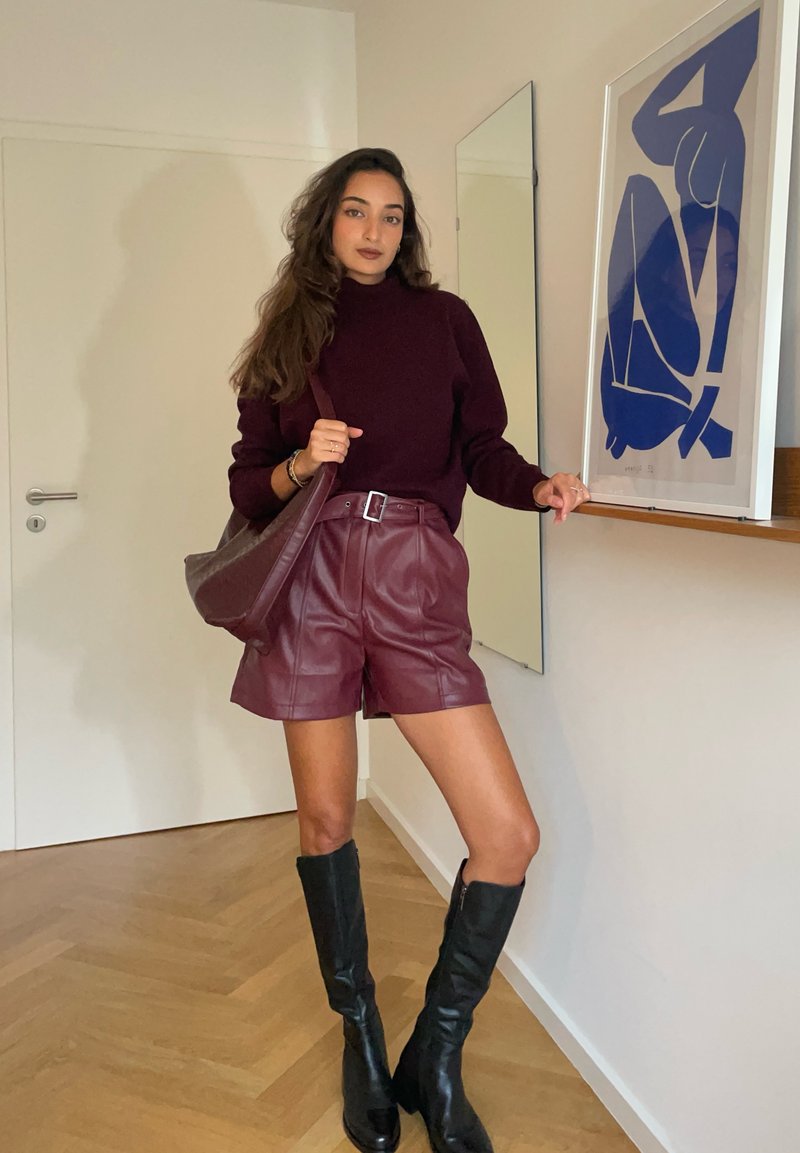 Burgunderfarbener Rollkragenpullover kombiniert mit hochtaillierten Leder Shorts. Schwarze kniehohe Stiefel. Große braune Tasche. Holzboden und Spiegel sichtbar.
