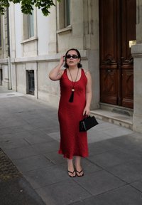 Donna in un abito rosso senza maniche, sandali neri, occhiali da sole e con una borsa nera in mano, che sta in un marciapiede cittadino vicino all'ingresso di un edificio.