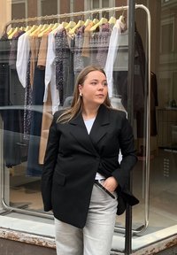 Črn suknjič s singlom gumbom, nosen čez belo srajco. Svetlo sivi hlače in velike biserne uhane. V ozadju so obešeni kosi oblačil.