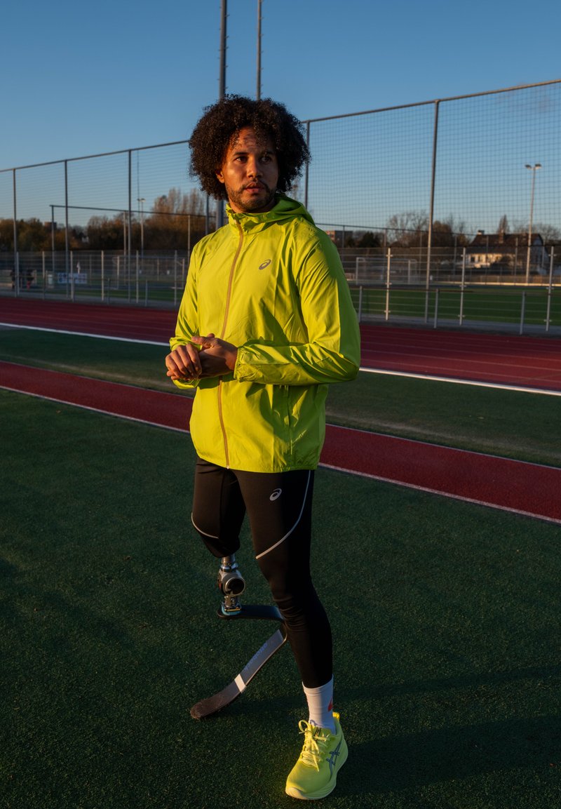 Atleta com um casaco leve amarelo vibrante, leggings desportivos pretos e sapatos amarelo néon, exibindo uma perna artificial com lâmina.