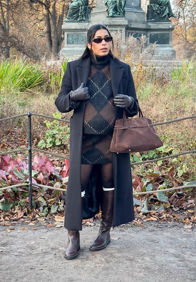 Abrigo negro sobre un vestido de punto marrón con estampado, botas marrones hasta la rodilla y guantes a juego. Bolso de ante marrón con asas cortas.