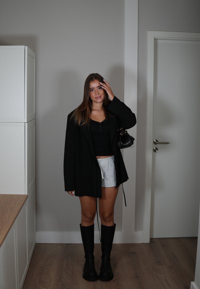 Blazer negro de gran tamaño sobre una blusa negra, pantalones cortos grises y botas altas negras. Sostiene un pequeño bolso negro. Suelo de madera y paredes lisas.