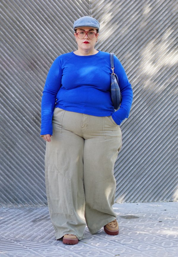 Una persona con una gorra azul y un jersey, pantalones beige anchos y un bolso al hombro está de pie frente a una pared metálica con nervaduras diagonales.